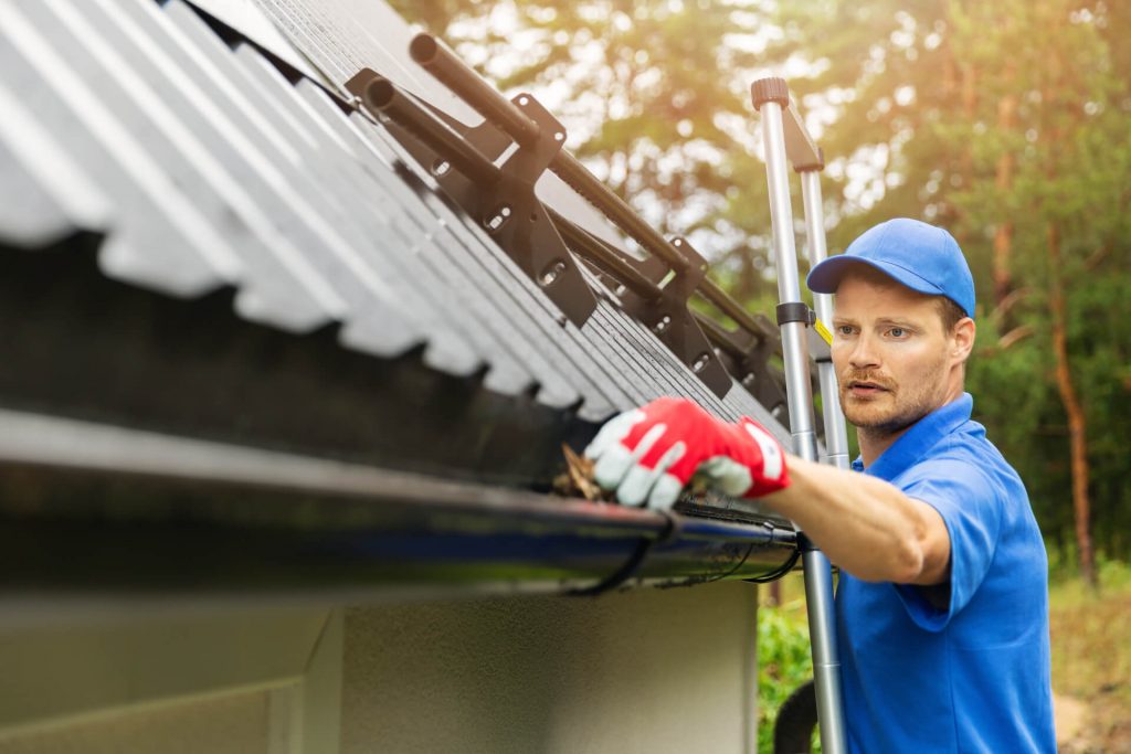 A man in a blue shirt and red gloves is cleaning out a gutter, focused on removing debris.